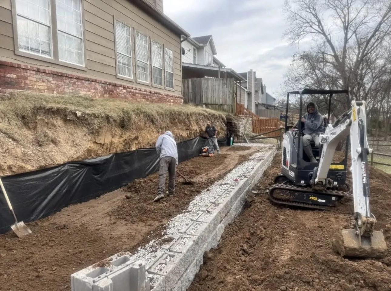 Mini excavator and crew setting block for an engineered 11-foot retaining wall with a French drain in Broken Arrow